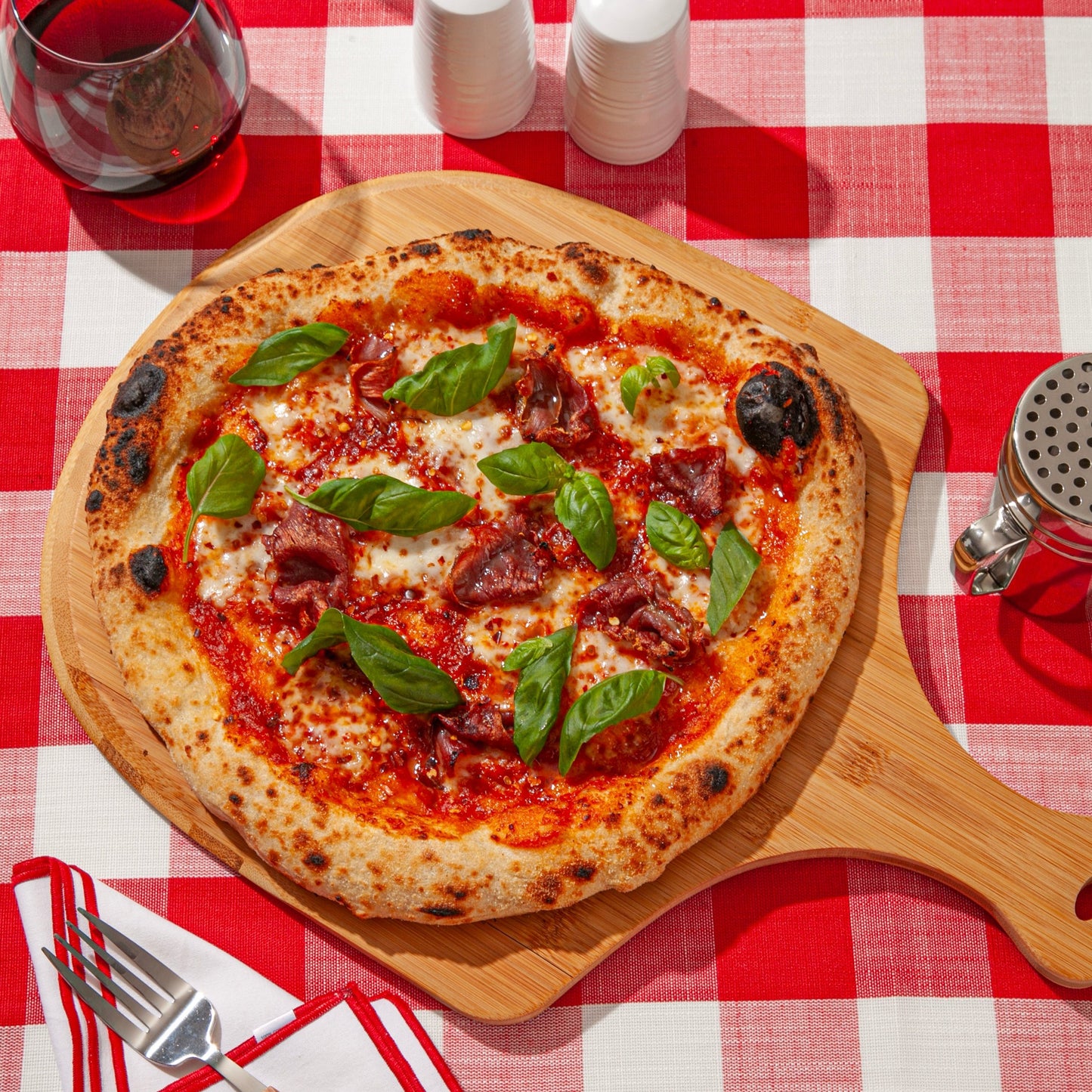 A wood-fired pizza with a crispy, charred crust, topped with tomato sauce, melted cheese, slices of cured meat, and fresh basil leaves. The pizza is served on a wooden board placed on a red and white checkered tablecloth, accompanied by a glass of red wine, a grated cheese shaker, and a neatly folded napkin with a fork and knife.