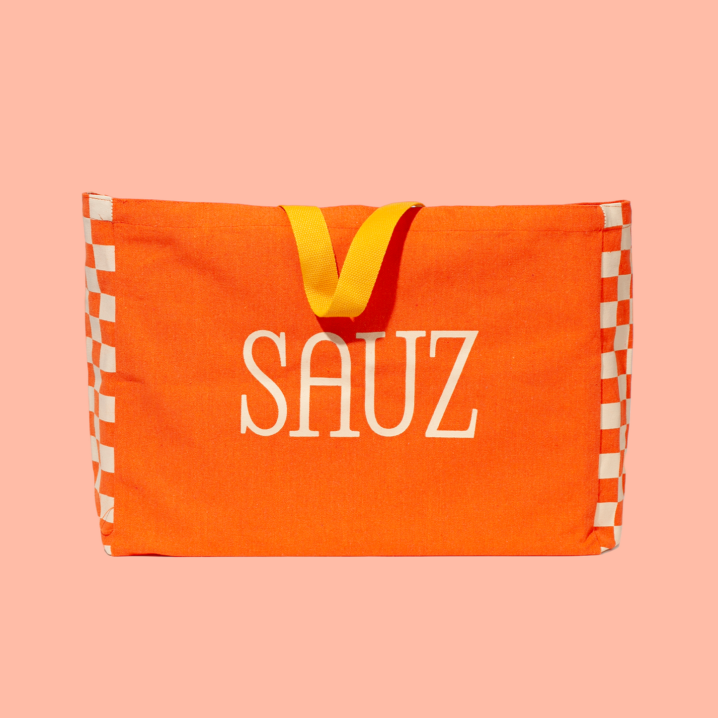 Bright orange tote bag with bold white 'SAUZ' text in the center, accented by yellow handles and a checkered pattern on the sides, set against a soft pink background