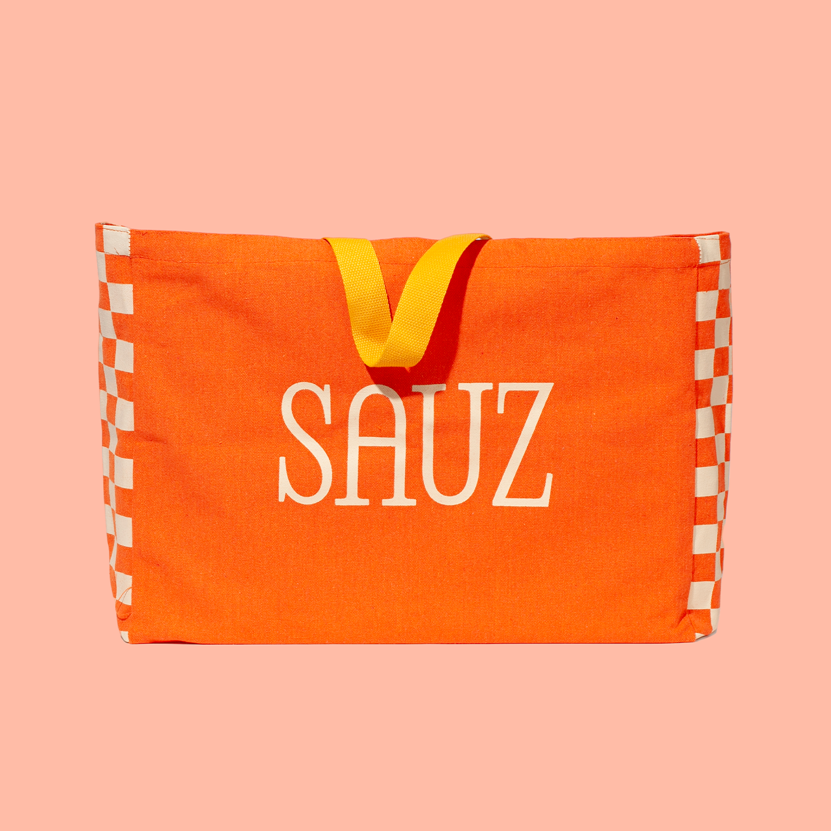Bright orange tote bag with bold white 'SAUZ' text in the center, accented by yellow handles and a checkered pattern on the sides, set against a soft pink background