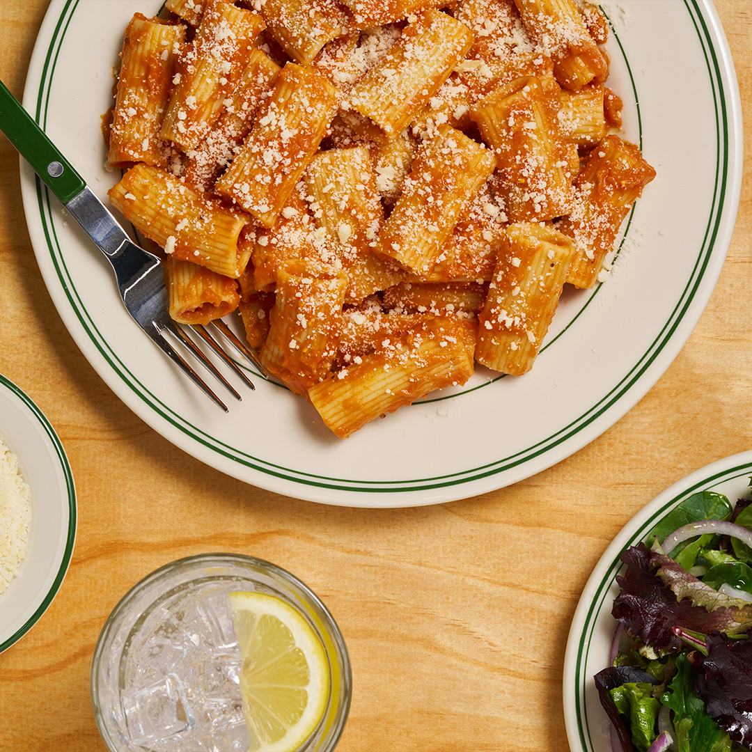 A plate of rigatoni pasta coated in a rich orange sauce and topped with a generous sprinkle of grated Parmesan cheese. The dish is served on a white plate with green rim accents, accompanied by a glass of ice water with a lemon slice, a side of fresh salad with mixed greens and red onion, and a small bowl of extra grated Parmesan on a wooden table