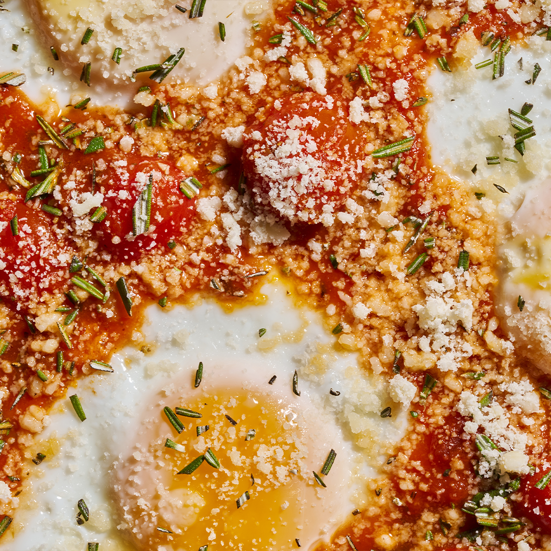 Close-up of baked eggs in a vibrant tomato sauce, garnished with fresh rosemary, grated cheese, and cherry tomatoes for a flavorful, textured dish