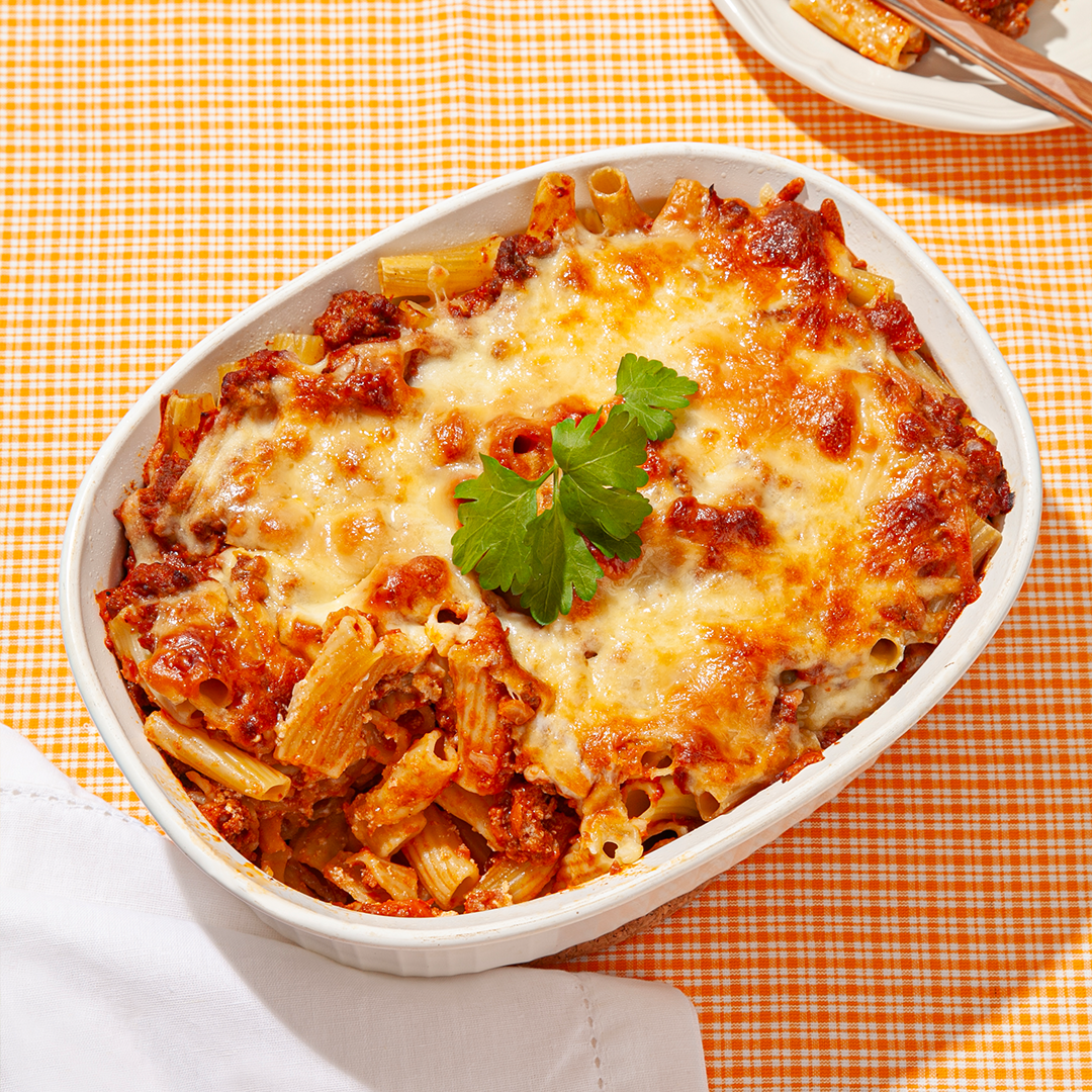 A baked ziti dish topped with golden, melted cheese and garnished with a sprig of fresh parsley, served in a white casserole dish on an orange gingham tablecloth, accompanied by a neatly folded white napkin