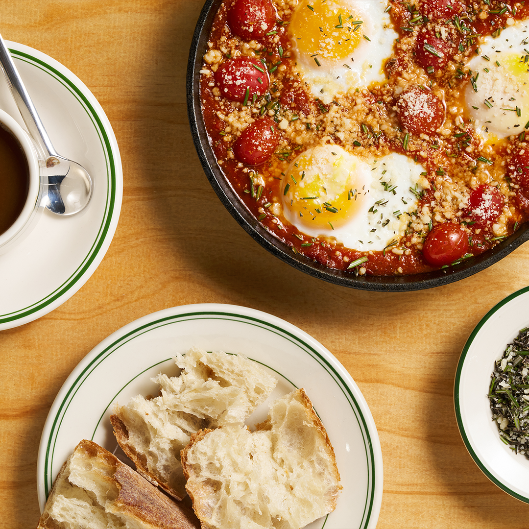 A hearty breakfast dish featuring baked eggs in a rich tomato sauce topped with herbs and cheese, served with fresh bread on the side and a cup of coffee, set on a warm wooden table