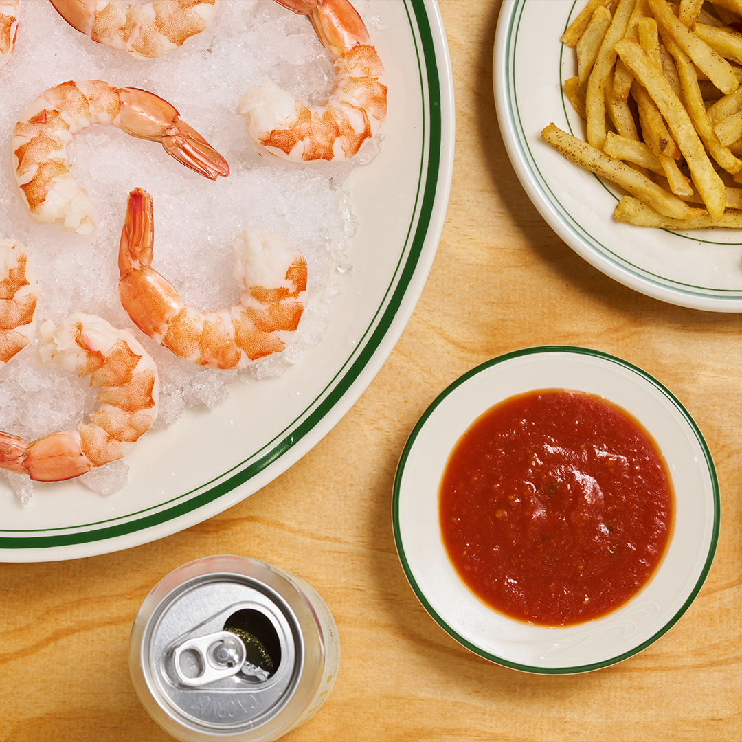 A plate of chilled shrimp served over crushed ice, accompanied by a bowl of red cocktail sauce, a side of seasoned fries, and a beverage can, arranged on a wooden table for a classic seafood appetizer presentation