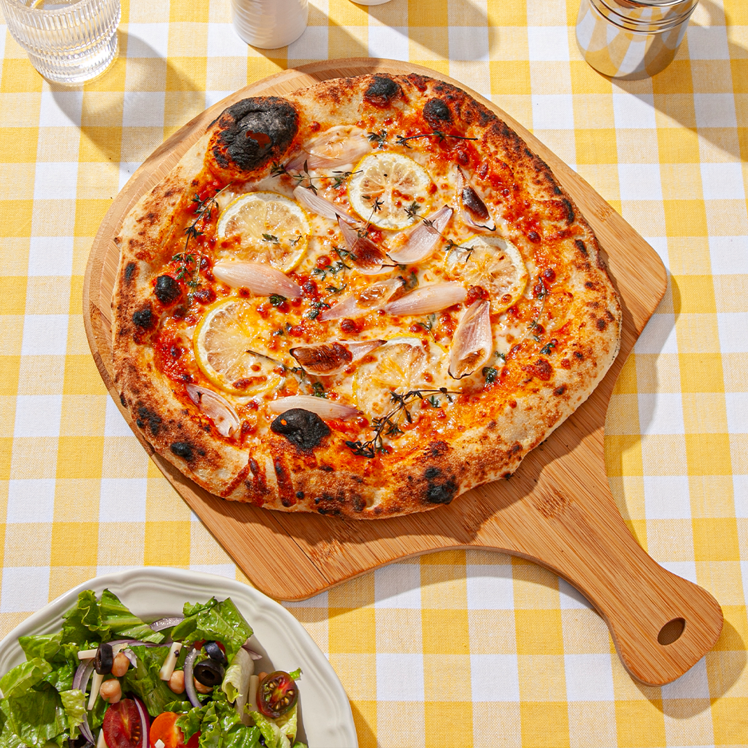 A wood-fired pizza topped with roasted lemon slices, caramelized shallots, fresh thyme, and a rich tomato sauce, presented on a wooden pizza peel over a yellow and white checkered tablecloth, accompanied by a crisp garden salad and a glass of water