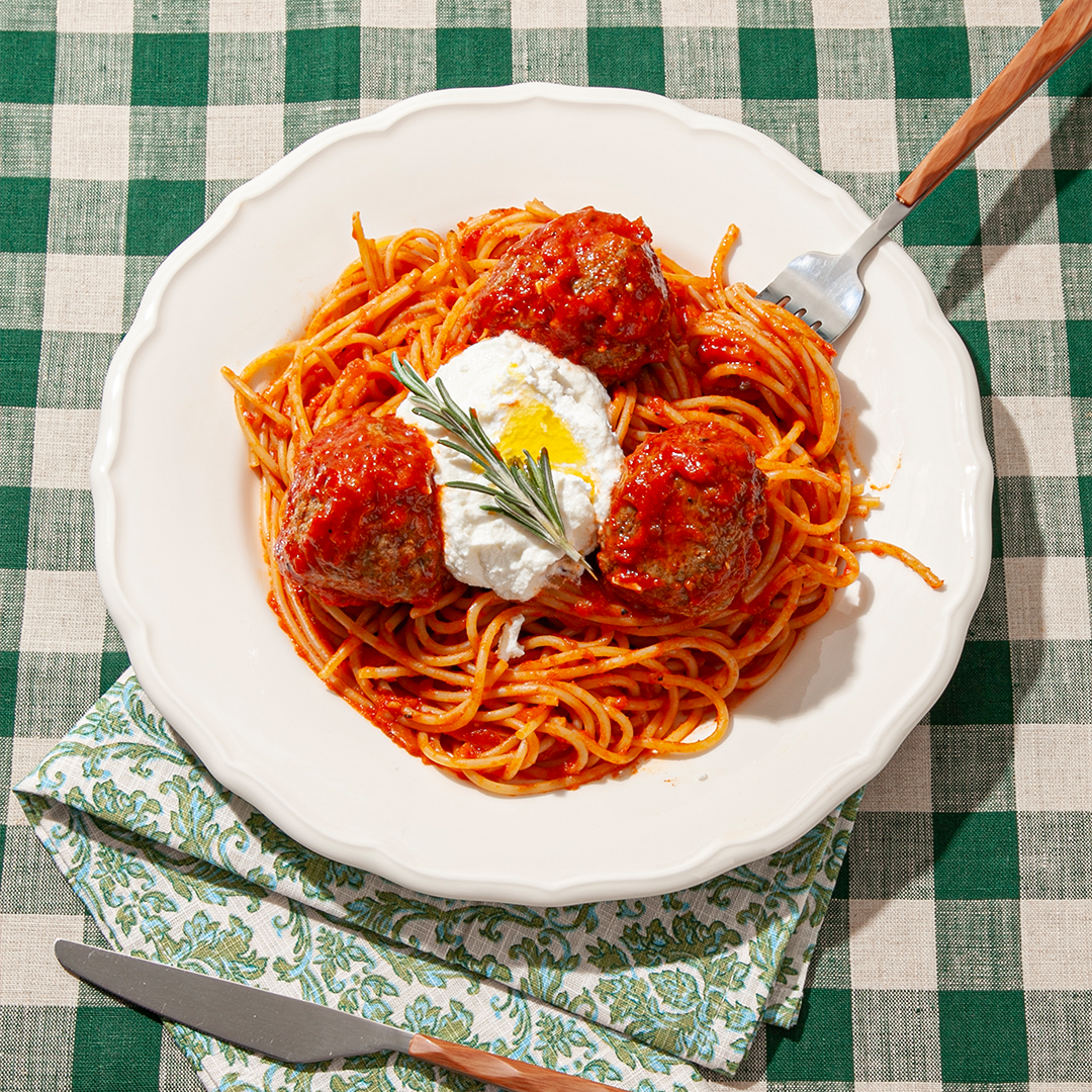 A plate of spaghetti topped with red tomato sauce, three meatballs, a dollop of ricotta cheese, a drizzle of olive oil, and a sprig of fresh rosemary. The dish is served on a white plate placed on a green and white checkered tablecloth with a patterned napkin underneath, accompanied by a fork and knife