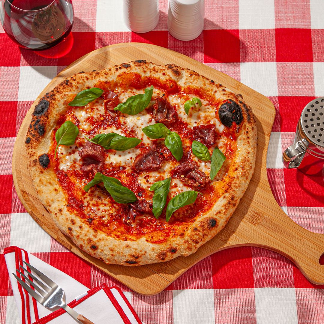 A freshly baked pizza topped with rich tomato sauce, mozzarella, fresh basil leaves, and slices of cured meat, presented on a wooden pizza peel over a red and white checkered tablecloth, accompanied by a glass of red wine and traditional pizza condiments