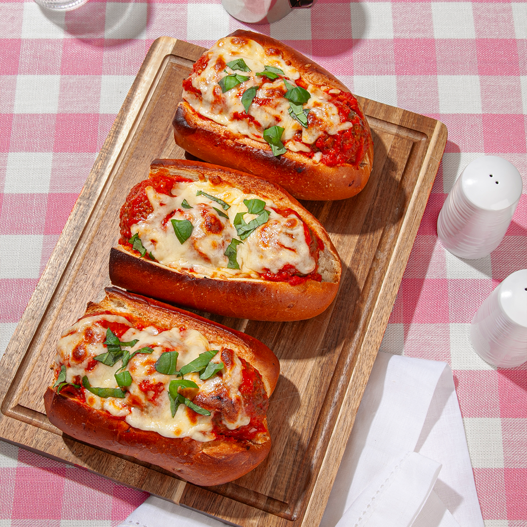 Three meatball subs topped with melted cheese and fresh basil, served on a wooden board with a pink and white checkered tablecloth background, alongside white salt and pepper shakers