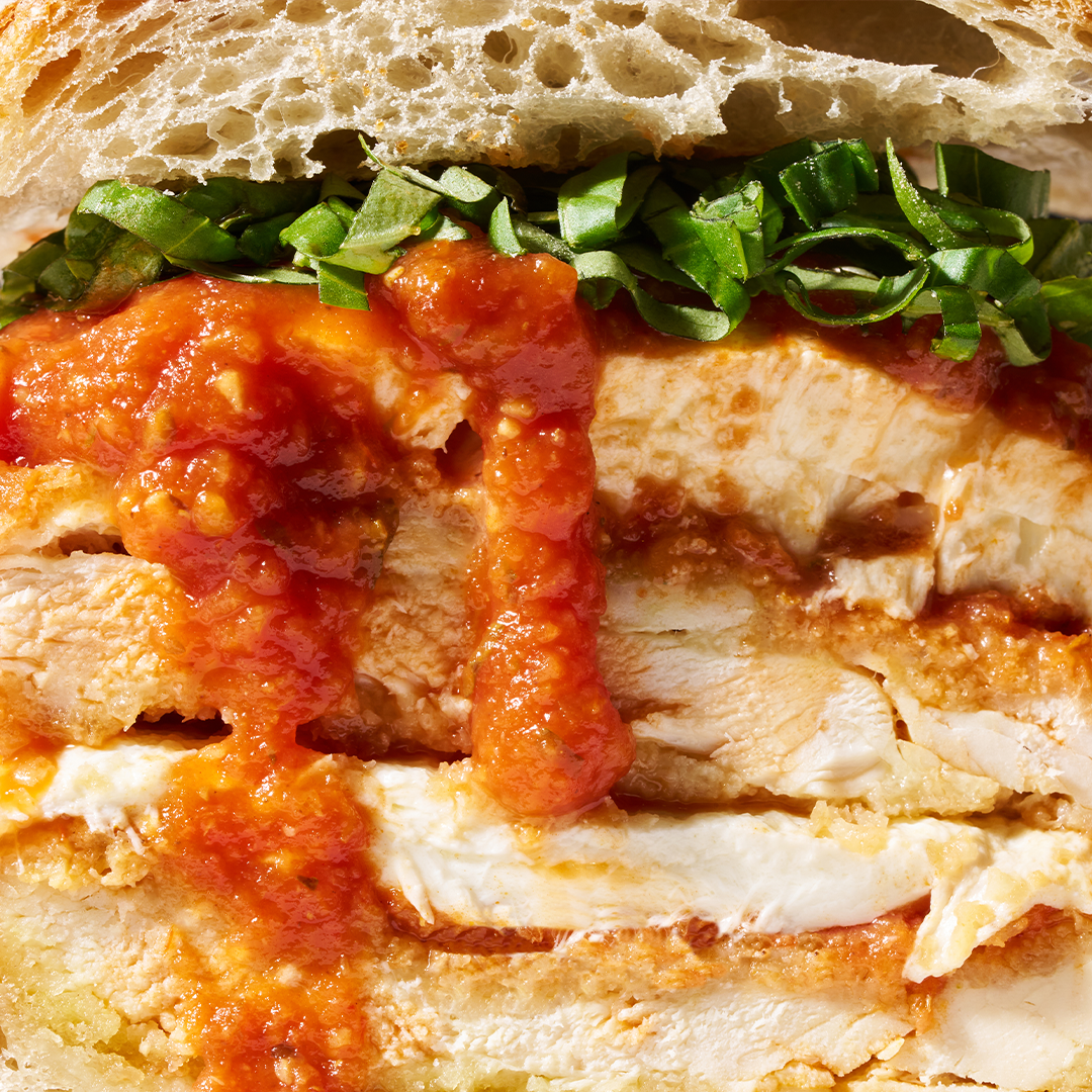 Close-up of a chicken parmesan sandwich with layers of breaded chicken, marinara sauce, melted cheese, and fresh basil, served on crusty bread