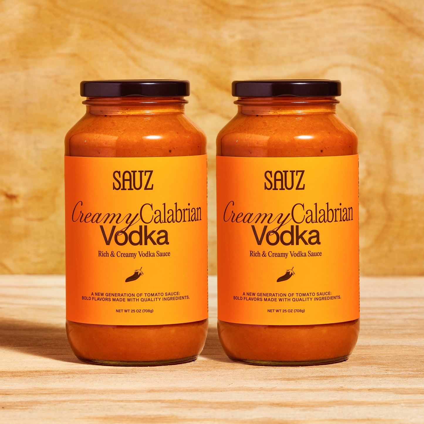 A set of two jars of Sauz Creamy Calabrian Vodka sauce, neatly lined up against a light wooden background. Each jar features a bold orange label with black text reading 'Rich & Creamy Vodka Sauce' and a small chili graphic. The jars contain creamy orange-red tomato sauce and are topped with black lids. The text highlights the sauce as 'A new generation of tomato sauce: bold flavors made with quality ingredients.' Each jar is labeled as 25 oz (708g).