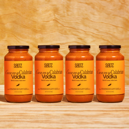 A set of four jars of Sauz Creamy Calabrian Vodka sauce, neatly lined up against a light wooden background. Each jar features a bold orange label with black text reading 'Rich & Creamy Vodka Sauce' and a small chili graphic. The jars contain creamy orange-red tomato sauce and are topped with black lids. The text highlights the sauce as 'A new generation of tomato sauce: bold flavors made with quality ingredients.' Each jar is labeled as 25 oz (708g).