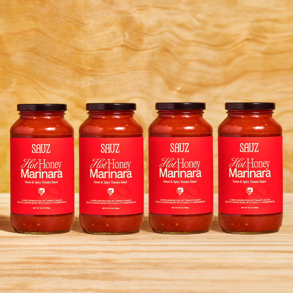 Four jars of Sauz Hot Honey Marinara arranged in a row on a wooden surface. Each jar features a bold red label with white text that reads 'Sweet & Spicy Tomato Sauce,' accompanied by a small honeycomb graphic. The jars are filled with vibrant red tomato sauce and sealed with black lids, set against a natural wooden background