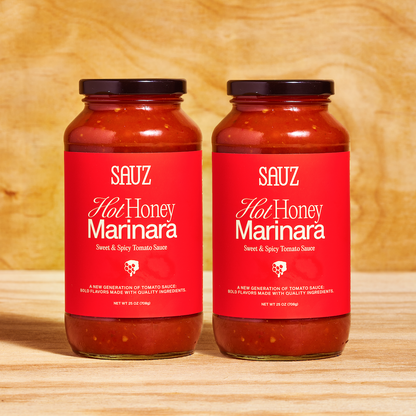 Two jars of Sauz Hot Honey Marinara placed side by side on a wooden surface. The jars feature vibrant red labels with white text reading 'Sweet & Spicy Tomato Sauce' and a small honeycomb graphic. Each jar contains rich red tomato sauce and is sealed with a black lid, displayed against a warm wooden background