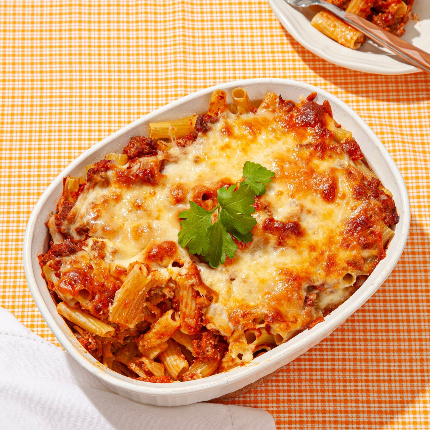 A baked pasta dish served in a white oval casserole dish, featuring rigatoni mixed with tomato sauce, ground meat, and melted golden-brown cheese. The dish is garnished with a sprig of fresh parsley and set on an orange gingham tablecloth, with a portion served on a smaller plate in the background