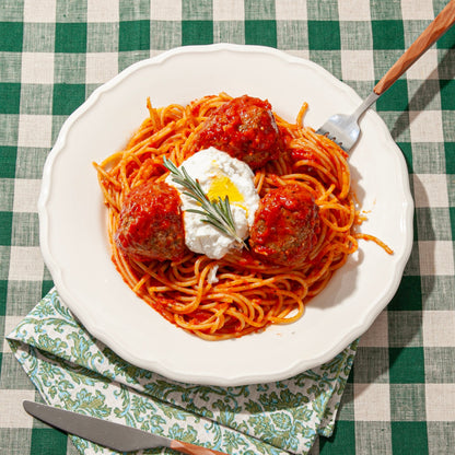 A plate of spaghetti topped with red tomato sauce, three meatballs, a dollop of ricotta cheese, a drizzle of olive oil, and a sprig of fresh rosemary. The dish is served on a white plate placed on a green and white checkered tablecloth with a patterned napkin underneath, accompanied by a fork and knife.