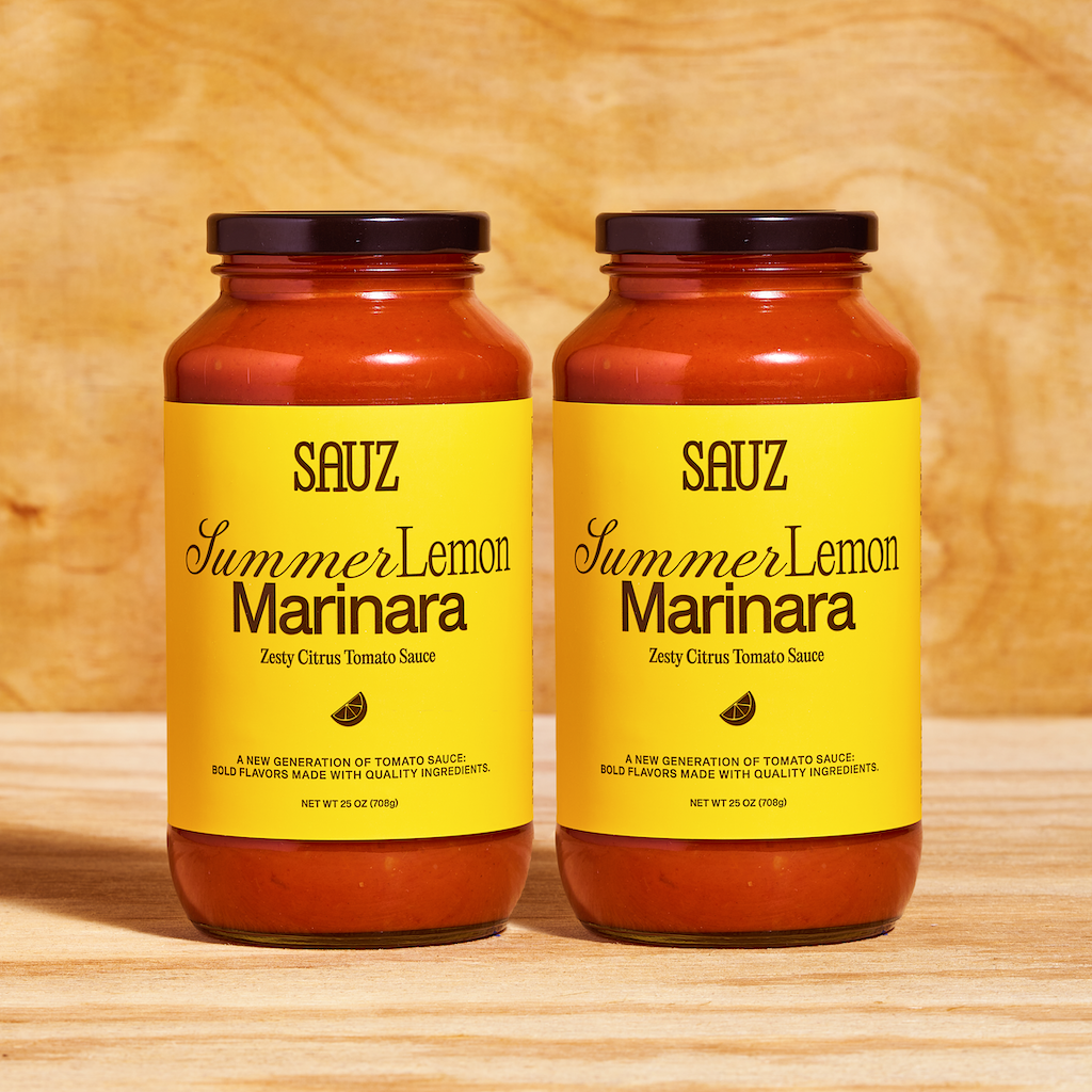 Two jars of Sauz Summer Lemon Marinara placed side by side on a wooden surface. The jars feature bright yellow labels with black text that reads 'Zesty Citrus Tomato Sauce,' accompanied by a small lemon wedge graphic. Each jar is filled with vibrant red tomato sauce and sealed with black lids, displayed against a natural wooden background