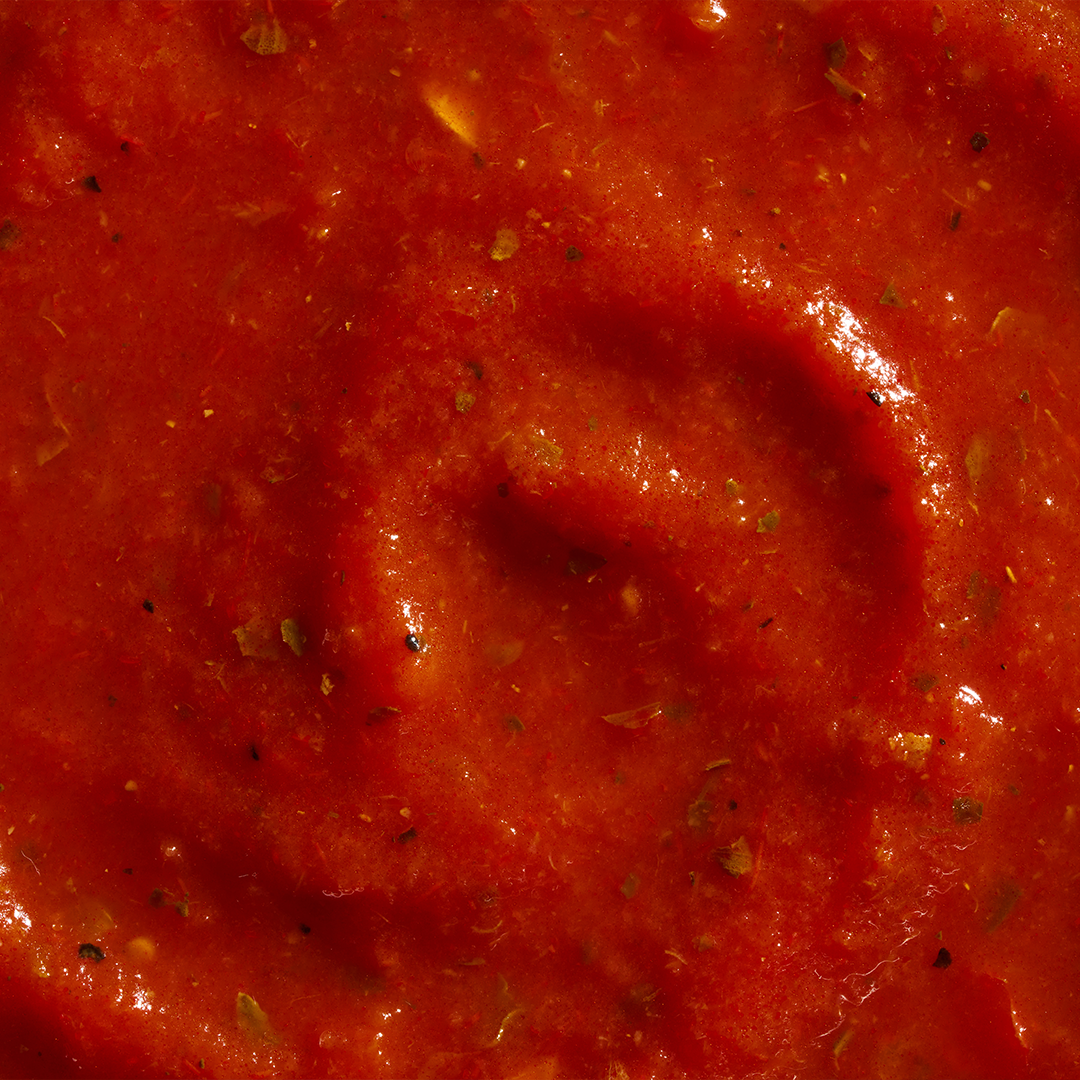 A close-up of bright red pasta sauce with a smooth yet slightly textured consistency, speckled with visible herbs and spices, showcasing a glossy finish