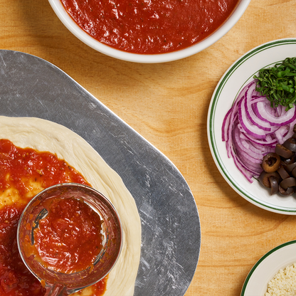 A pizza-making setup featuring dough on a metal peel being spread with tomato sauce using a ladle. A bowl of rich red tomato sauce sits nearby, along with a plate containing sliced red onions, black olives, and chopped herbs, and another plate with grated cheese, all arranged on a wooden surface